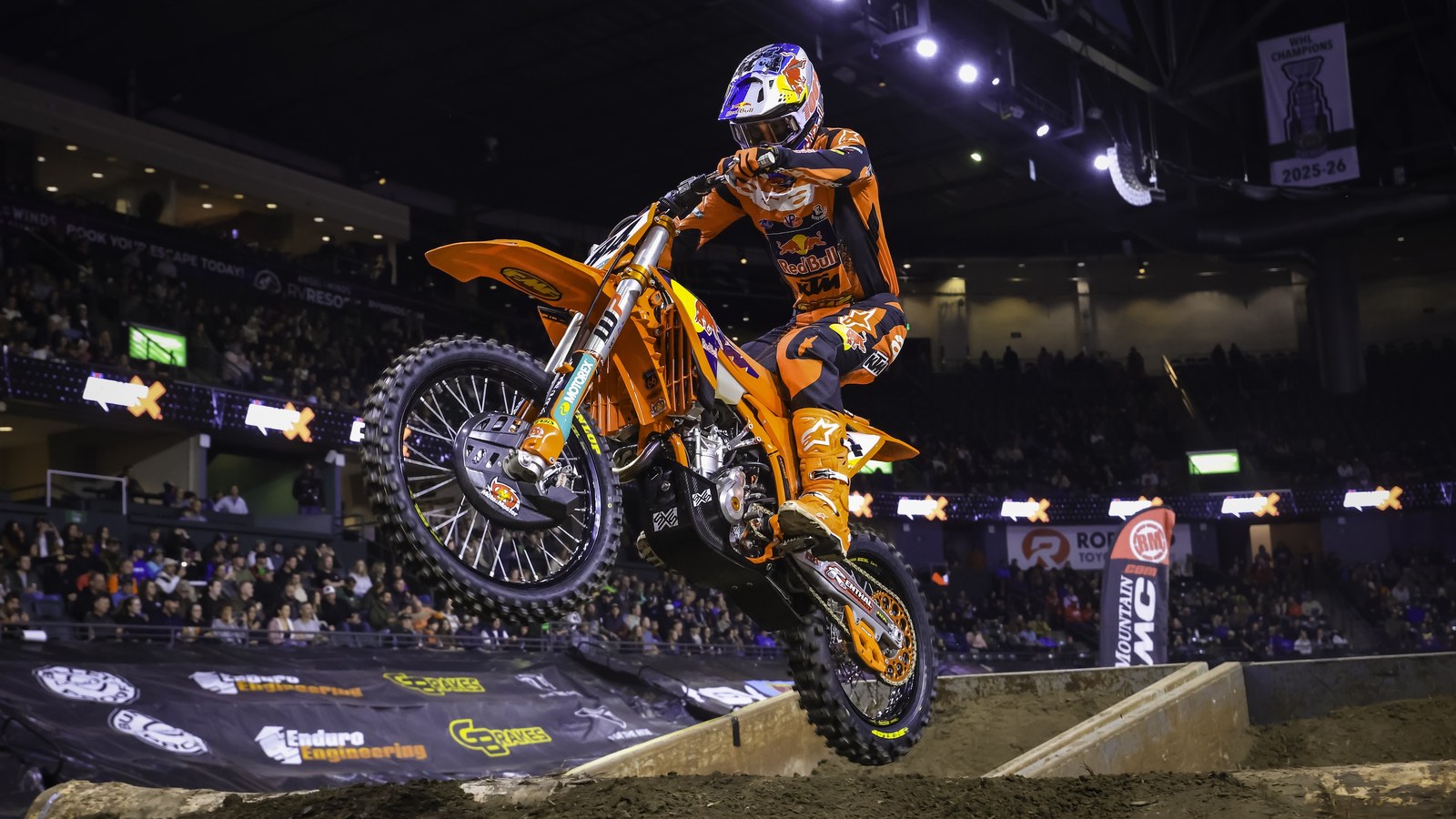 A rider in orange Red Bull KTM gear and a blue, red and yellow helmet jumps an orange KTM off a dirt step, with arena lights and fans in the stands behind.
