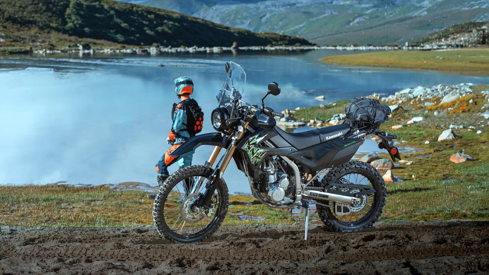 A black 2027 Kawasaki KLX300 XPL ABS with windshield and a bag strapped to its rear rack, parked beside a mountain lake while its rider looks out over the water