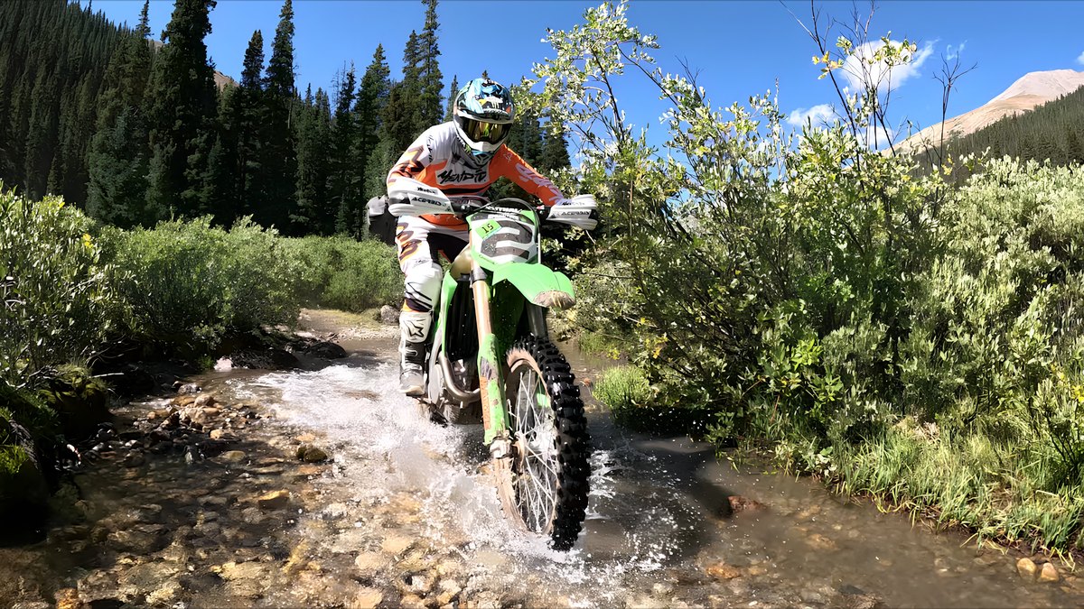 A Colorado 500 rider on a green dirt bike splashes through a mountain creek crossing