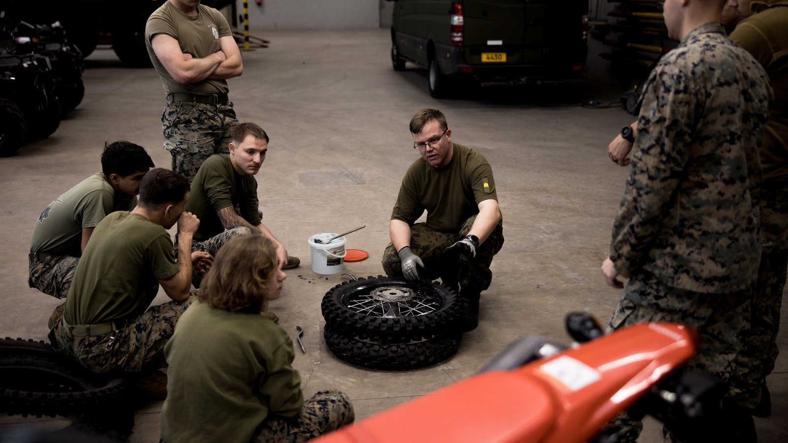 A man in gloves and glasses kneels on a garage floor beside two stacked spoked wheels with knobby tires, next to a white bucket with a brush and an orange lid, while people in olive shirts and camouflage pants watch. An orange dirt bike fender sits in the foreground.