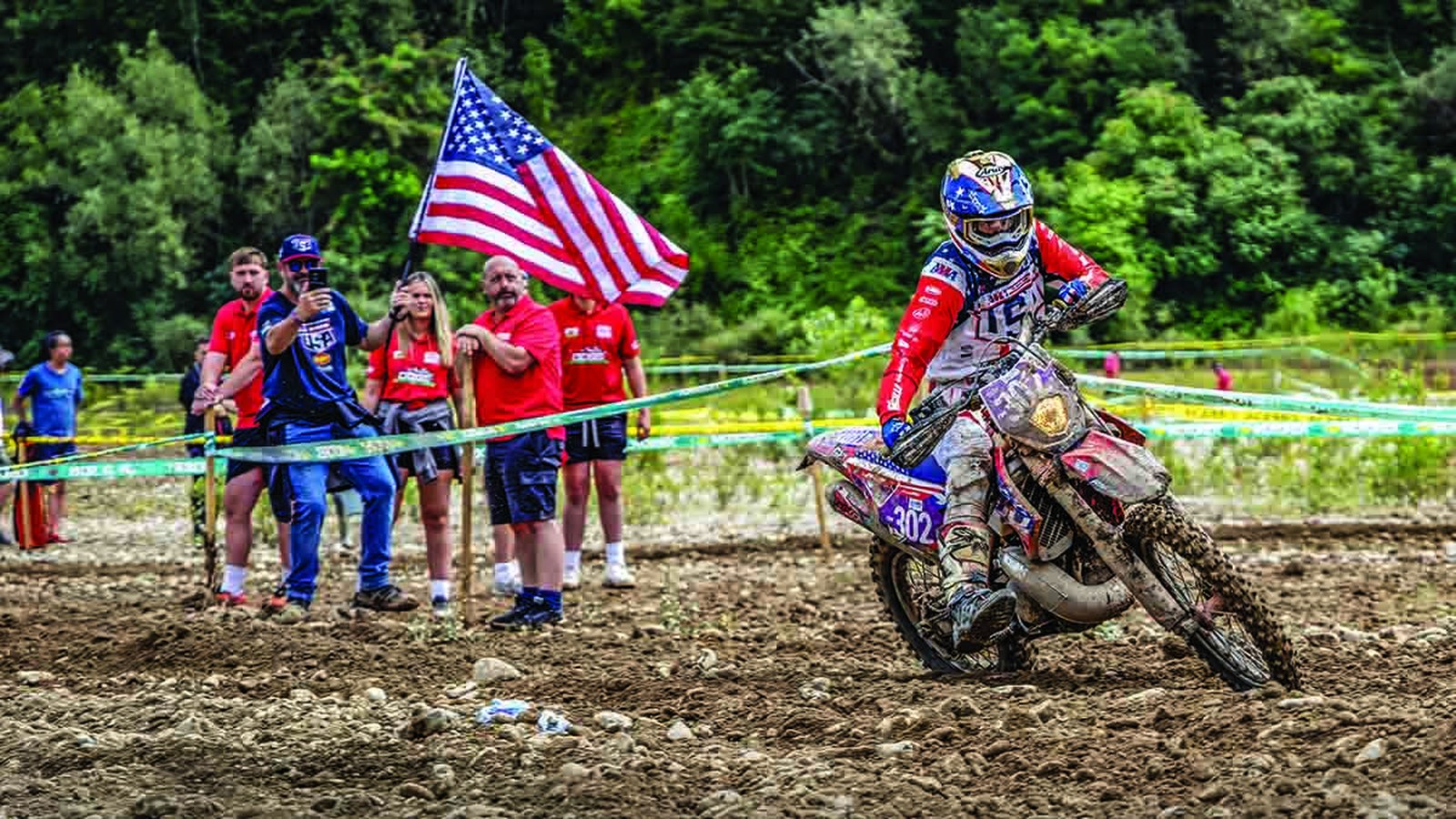 Enduro rider number 302 in red, white and blue kit on rocks, spectators with a US flag behind FIM tape