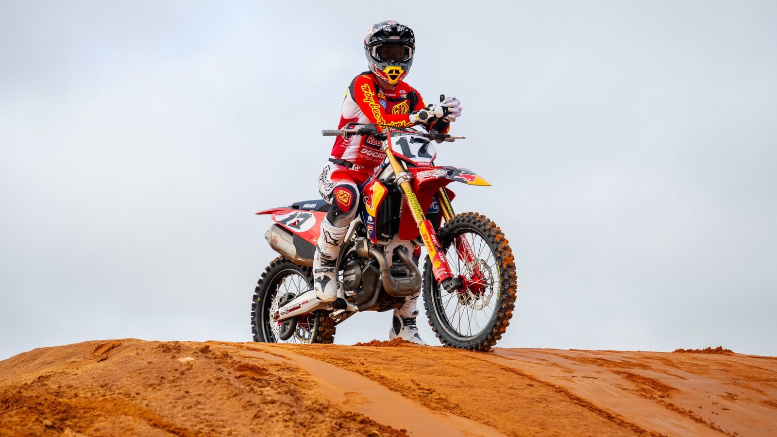 A rider in a red, white and yellow Troy Lee Designs jersey and black helmet sits on a red Ducati dirt bike with number 17 on the front plate, parked on top of an orange dirt mound under a gray sky