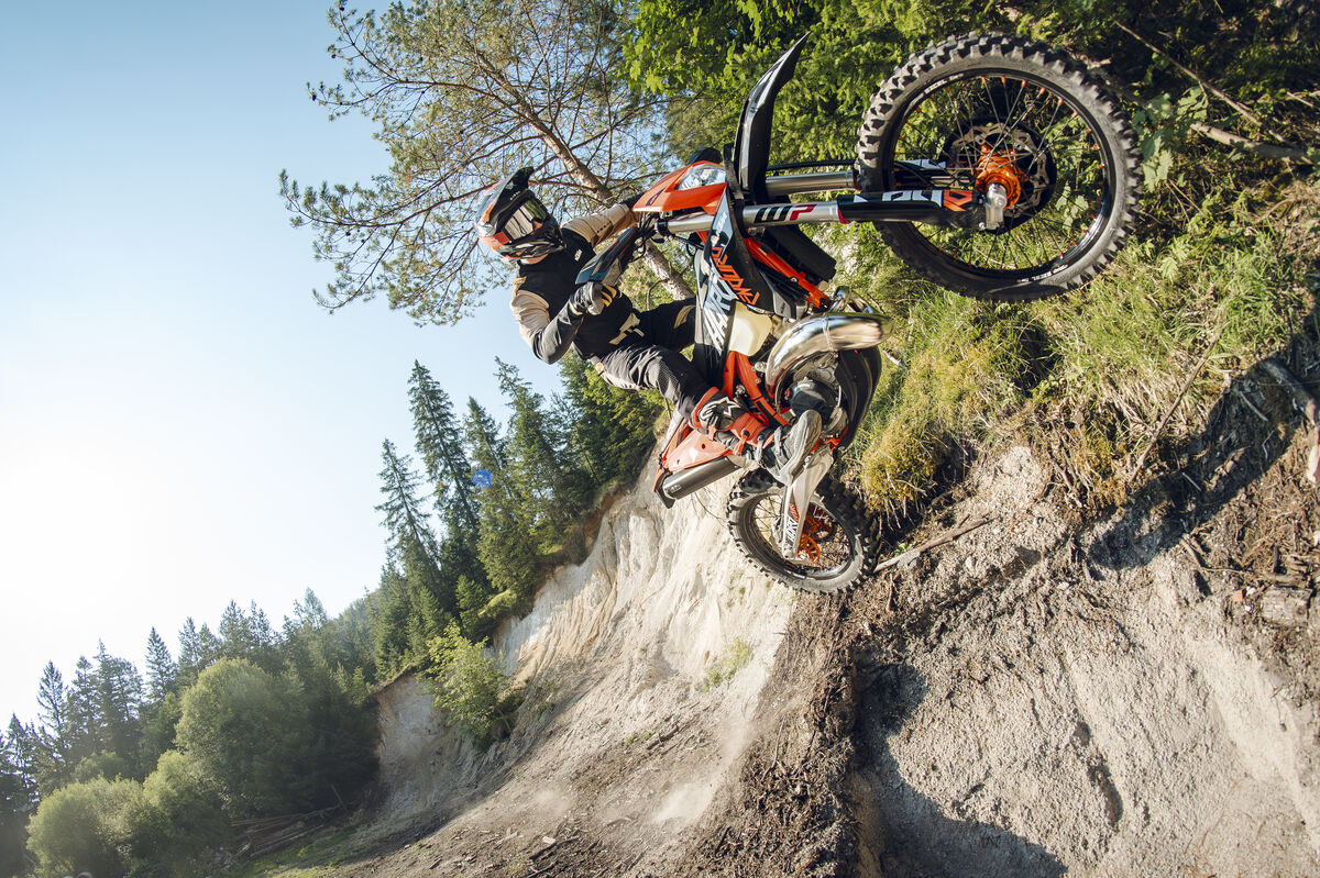 Rider wheelies an orange KTM up a steep, loose dirt bank below pine trees and blue sky