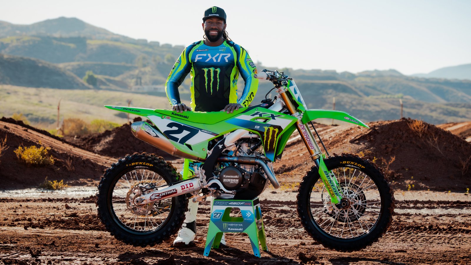 A smiling rider in a black Monster Energy cap and a blue, lime green and black FXR jersey stands behind a green Kawasaki dirt bike with number 27 on the side plate, parked on a stand on a dirt track with brown hills behind