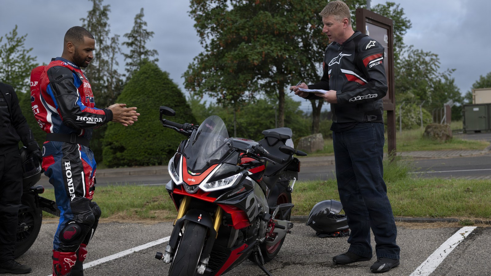 Two riders, one in a red, white and blue Honda race suit and one in a black leather jacket holding papers, talk beside a red and black Aprilia sport bike in a parking lot.