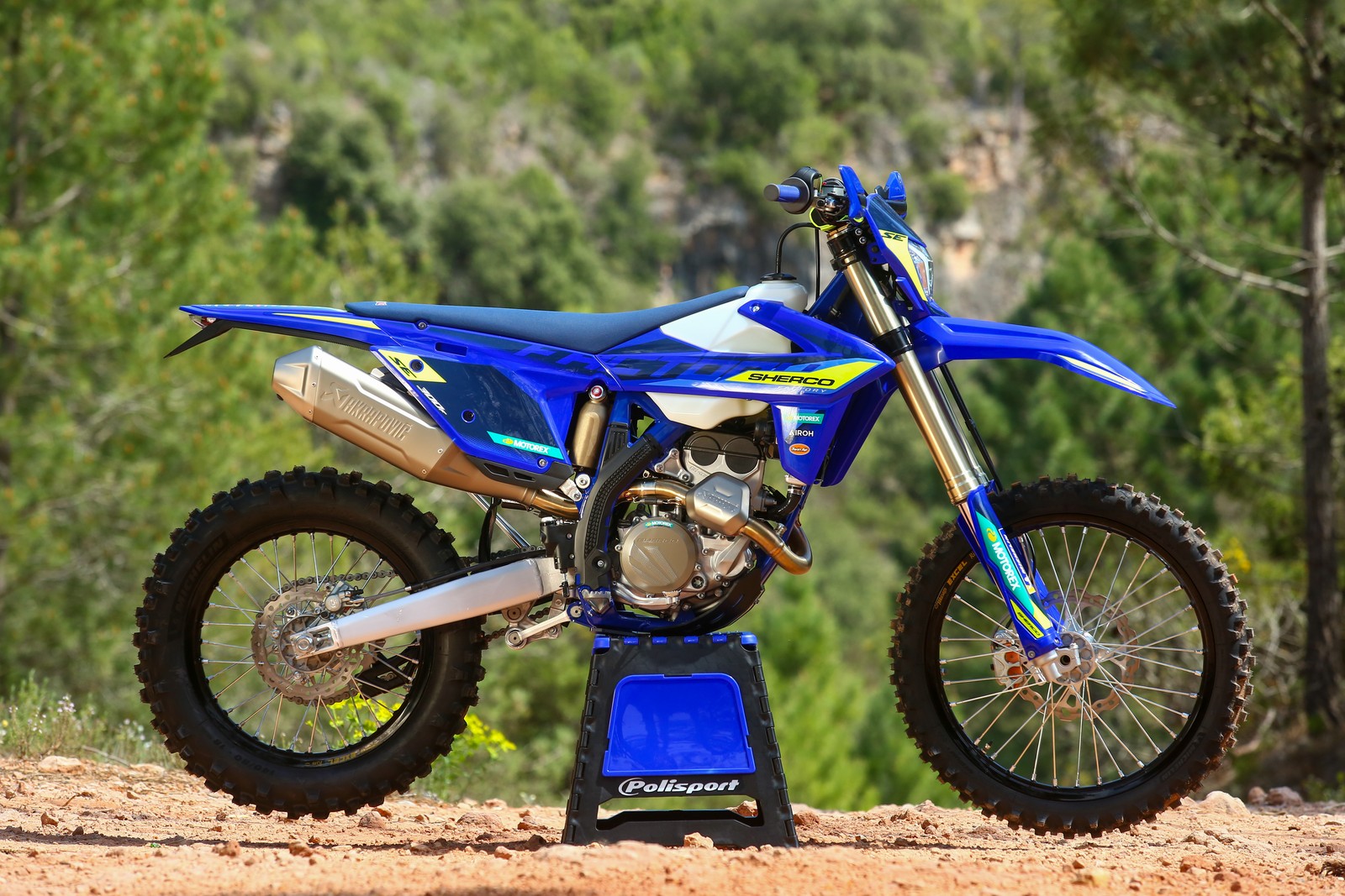 Blue 2027 Sherco 300 SEF Factory with an Akrapovič muffler, right-side view on a Polisport stand with pine forest behind